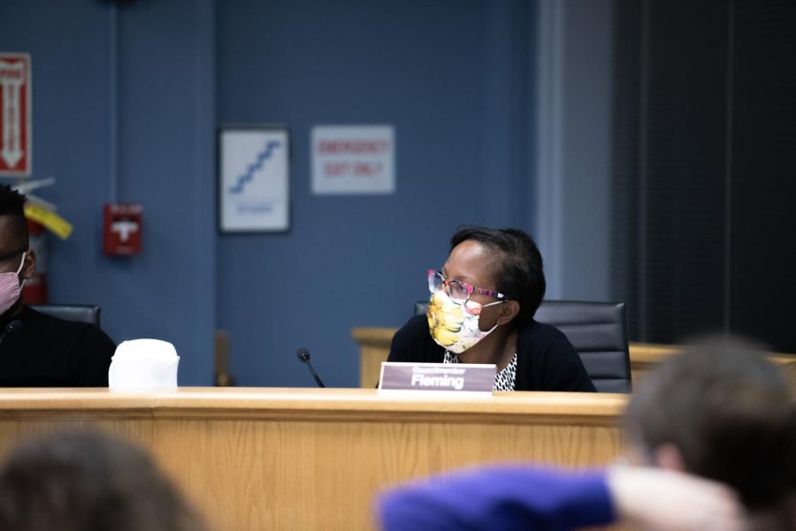 Cicely Fleming sits at her desk at City Council. The desk is brown, the background is blue and the chair is black. She is wearing a face mask.