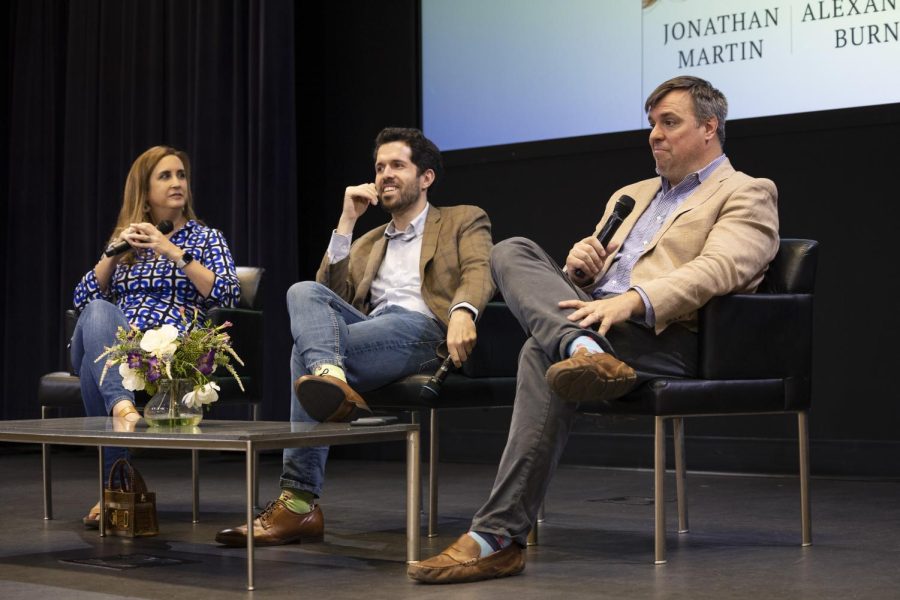 Three people dressed formally, sitting in chairs and holding microphones with a coffee table holding a flower vase in front of them.